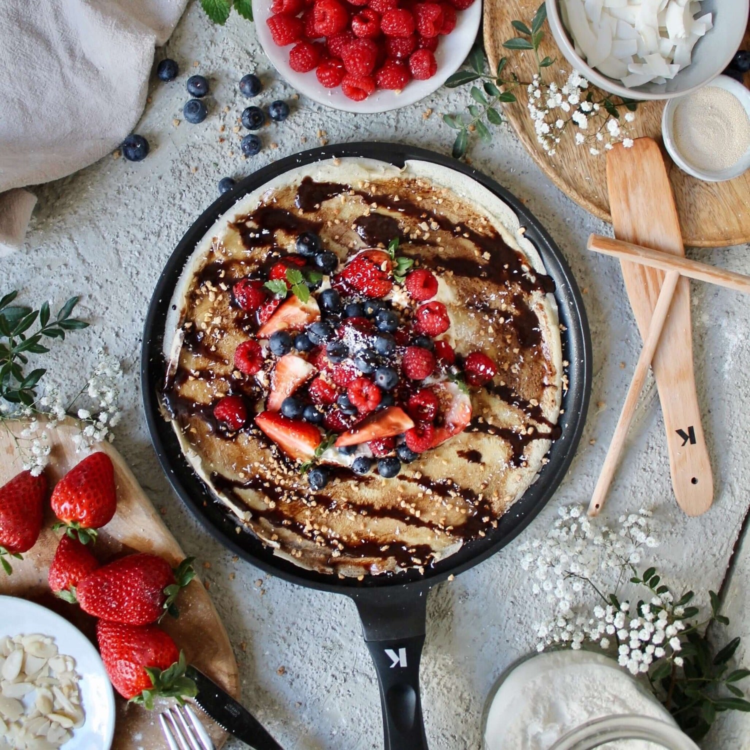 Crêpespfanne 28cm filled with mixed berries and chocolate sauce, surrounded by fresh fruit and toppings on a rustic table.