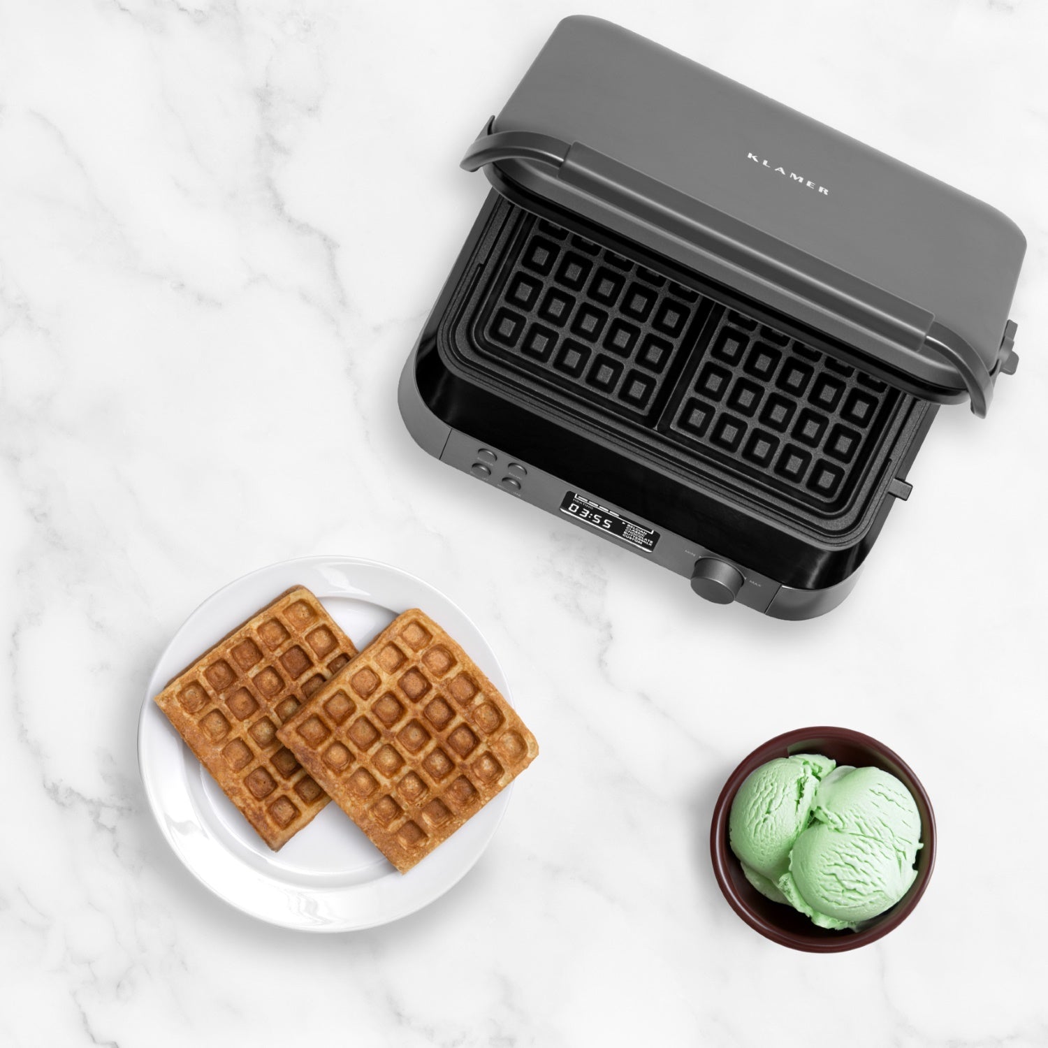 Waffeleisen mit zwei frisch gebackenen Waffeln und einer Schale mit grüner Eiscreme auf einem Marmortisch.