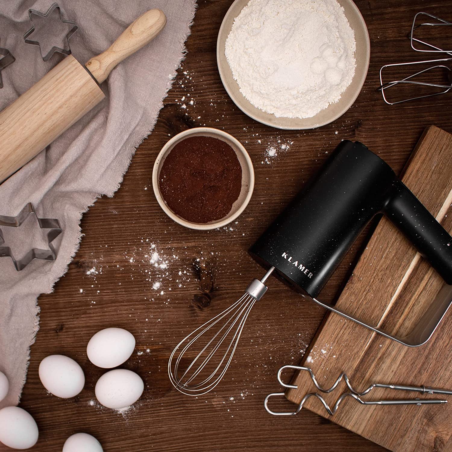 Handmixer with whisk, flour, cocoa powder, eggs, and baking tools on wooden table.